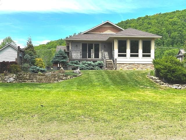 Rear view of the cottage from across the lawn, showing the bank of sunroom windows, a small deck, and the wooded ridge rising behind the roofline
