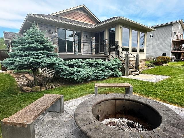 A second read of the rear yard: paver patio with a circular stone fire ring in the foreground, wood-railed deck beyond, blue spruces and the rear of the cottage in the distance