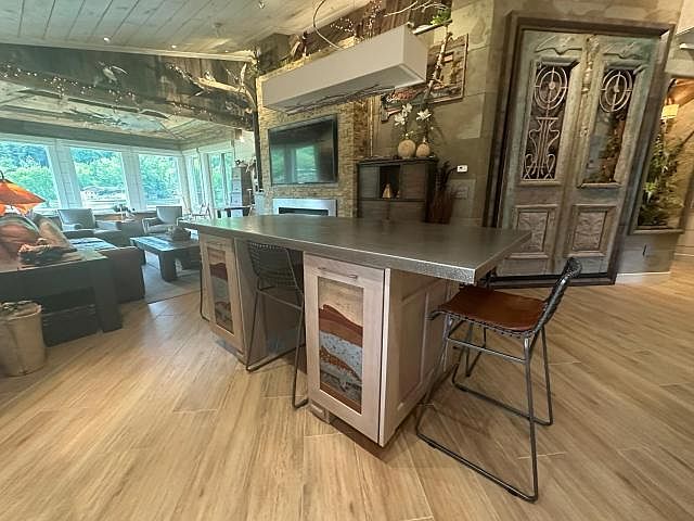 Tight interior view of the great room: the wrought-iron antique doors on the right wall, the kitchen island with its concrete-look top, the fireplace and mounted TV behind, sunroom windows opening to the trees beyond